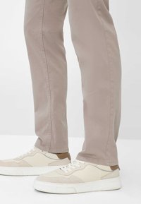 Helle beigefarbene Baumwollhose mit Slim Fit und geraden Beinen, kombiniert mit weißen Sneakers und einem braunen Akzent auf den Schuhen.