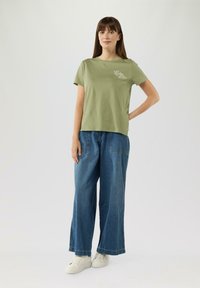 Olijfgroene T-shirt met korte mouwen met witte tekst; gecombineerd met een brede blauwe denim broek en witte sneakers. Eenvoudig ontwerp.