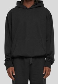 Felpa nera oversize realizzata in tessuto morbido, con tasca frontale, cappuccio con coulisse e polsini a coste, abbinata a pantaloni coordinati.