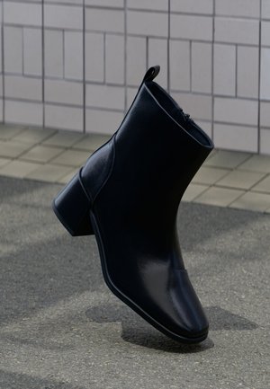Bottine en cuir noir avec talon bloc, présentée debout sur un pavé texturé avec un mur carrelé en arrière-plan.