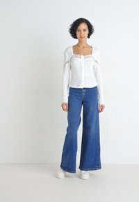Witte geribde top met lange mouwen, knoopsluiting en kanten accenten, gecombineerd met blauwe wijde pijpen jeans en witte sneakers.