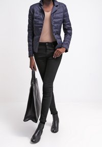 Marinblå vadderad jacka med en figursydd design, beige topp, svarta skinny jeans, ankelskor och en svart tote-väska. Slät textur, knapppdetaljer.