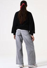 Suéter negro de gran tamaño con mangas abullonadas, combinado con pantalones de denim gris holgados. Es visible una cinta roja en el pelo largo y ondulado.