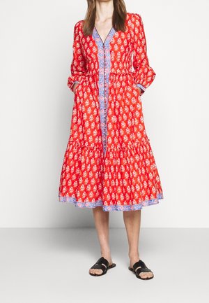 Femme portant une robe midi rouge à fleurs avec bordure bleue, boutonnée sur le devant, manches longues et sandales noires à bouts ouverts, debout les mains dans les poches.