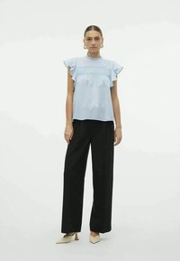 Blusa azzurra chiara con maniche a volant e dettagli smock, abbinata a pantaloni larghi neri e scarpe con tacco neutre.