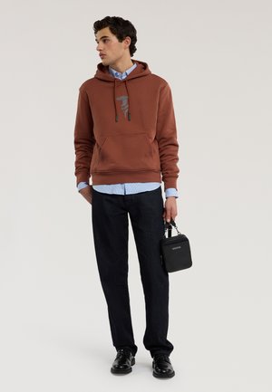 Joven con sudadera marrón sobre una camisa azul, pantalones negros, zapatos negros, sosteniendo una pequeña bolsa negra en su mano derecha, de pie frente a un fondo liso.