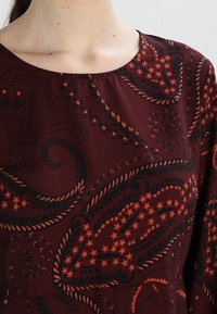 Robe en tissu bordeaux avec un motif paisley noir et rouge, texture lisse, encolure ronde et manches longues.