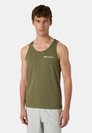 Giovane uomo con canotta verde oliva Champion e pantaloncini grigio chiaro in piedi contro uno sfondo semplice.