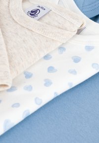 Tre capi leggeri sovrapposti: uno beige con una finitura testurizzata, uno bianco con motivi a cuore blu e uno blu pastello uniforme.