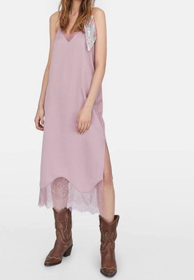Rosa ärmelloses Kleid mit V-Ausschnitt, mit einer Spitze am Saum und einem seitlichen Schlitz. Akzentuiert mit einem dekorativen Detail an der linken Schulter. Braune Cowboystiefel.
