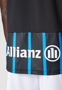 Camisola atlética preta com uma superfície texturizada, apresentando riscas verticais azuis e um logótipo branco "Allianz" na barra inferior.