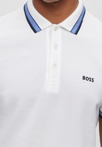 Polo blanc en tissu texturé, avec un col rayé bleu et noir, trois boutons et un logo "BOSS" brodé.