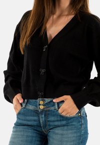 Cardigan lavorato a maglia nero con scollatura a V e bottoni decorativi a forma di fiocco, abbinato a jeans in denim azzurro con hardware color oro.