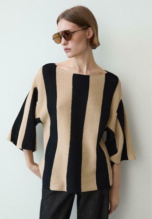 Frau mit kurzen Haaren, die große braune Sonnenbrillen und einen locker gestrickten Pullover mit breiten schwarzen und beigen vertikalen Streifen trägt.