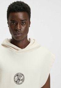 Beige mouwloze hoodie met een capuchon, voorzien van een cirkelvormig zwart grafisch logo op de borst. Het materiaal lijkt zacht en gestructureerd.