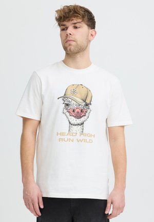 Mann med hvit T-skjorte med strutsehode i beige caps og tekst "HEAD HIGH RUN WILD" på ensfarget lys bakgrunn.