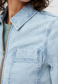 Hellblaues Denim-Jackett mit spitzem Kragen, einer Vordertasche und einem silbernen Reißverschluss. Der Stoff hat eine strukturierte Oberfläche mit subtilen Variationen.