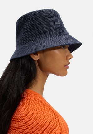 Kobieta z długimi ciemnymi włosami, w teksturowanym granatowym kapeluszu typu bucket oraz pomarańczowym dzianinowym swetrze, zwrócona w prawo na jednolitym tle.
