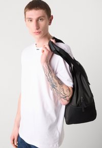 Černý batoh s hladkým povrchem, vybavený zipovou kapsou a pevnými popruhy, držený osobou vebělicí tričko.