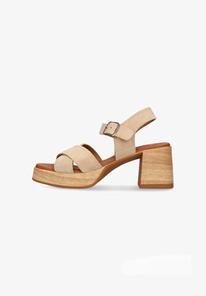 Sandale en daim beige avec talon bloc en bois, bride à la cheville et fermeture par boucle, présentant des brides croisées à l’avant-pied et une semelle intérieure rembourrée.