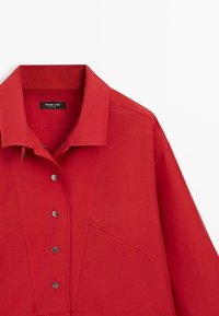 Chemise rouge à manches longues avec col pointu, cinq boutons-pression métalliques sur le devant et détails de coutures visibles sur la poitrine et les épaules.