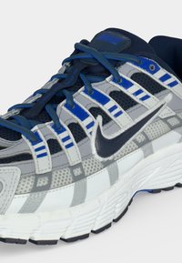 Λευκό και γκρι παπούτσι για τρέξιμο Nike με μπλε λεπτομέρειες, μπλε κορδόνια και μαύρο λογότυπο Nike στη πλευρά.
