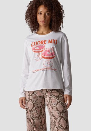 Frau mit lockigem Haar trägt ein weißes Langarmshirt mit roter Grafik von Trinkgläsern und Text, kombiniert mit pinken und braunen Schlangenprint-Hosen.