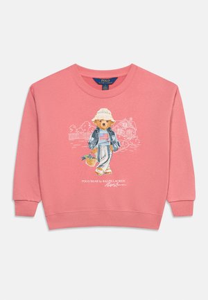 Sudadera rosa de manga larga con un oso estampado que lleva un sombrero, chaqueta, camisa de rayas, pantalones y sostiene una cesta, de pie frente a un boceto de una casa.
