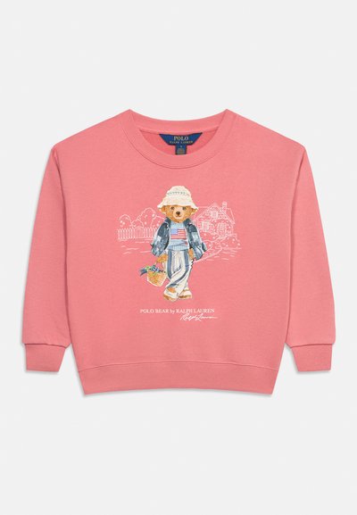 Ροζ μακρυμάνικη μπλούζα φούτερ με τυπωμένη αρκούδα που φοράει καπέλο, σακάκι, ριγέ πουκάμισο, παντελόνι και κρατάει ένα καλάθι, στέκεται μπροστά σε σχέδιο σπιτιού.