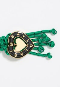 Heart-shaped accessory with black enamel and gold accents, featuring green braided cords and decorative knots with a patterned detail.