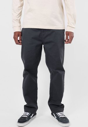 Homme portant un pantalon noir, une chemise blanche à manches longues et des baskets noires avec des lacets blancs, debout devant un fond clair.