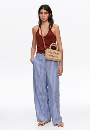 Mujer con top marrón de halter y pantalones sueltos azules a rayas, llevando un bolso cruzado tejido con la etiqueta BIMBA Y LOLA.