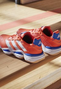 Červenooranžové sportovní boty s bílými pruhy, modrými akcenty, texturovaným materiálem a polstrovaným límcem, umístěné na dřevěném povrchu.