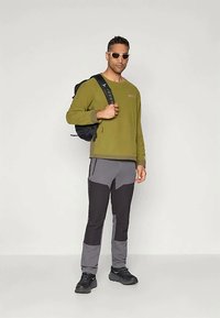 Maglietta a maniche lunghe verde oliva, pantaloni grigi e neri, zaino nero e occhiali da sole. Vestibilità atletica con pannelli di colore a contrasto e tasche.