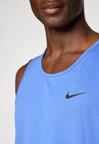 Mand iført en blå ærmeløs atletisk top med et sort Nike swoosh-logo på brystet, mod en ensfarvet hvid baggrund.
