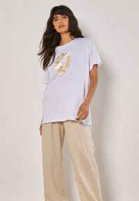 Weißes, übergroßes T-Shirt mit einem goldenen Grafikdesign auf der Vorderseite. Kombiniert mit hellbeigen weitem Hosen. Glatter Stoff und entspannte Passform.