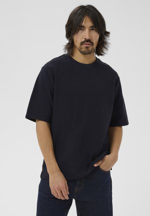 Homme aux cheveux mi-longs et à la moustache, portant une chemise noire texturée à manches courtes surdimensionnée et un jean bleu foncé, la main dans la poche.