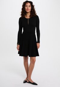 Schwarzes geripptes Kleid mit tailliertem Oberteil und Knopfdetails sowie einem weit schwingenden Faltenrock. Das Material ist weich und hat eine fließende Textur.