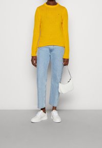 Pull en maille côtelée jaune, jean droit bleu clair, baskets blanches et petit sac matelassé blanc avec une bandoulière chaîne.