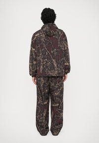 Giacca oversize con motivo camouflage e pantaloni abbinati in tonalità di marrone, bordeaux e nero, dotata di cappuccio e polsini elastici.