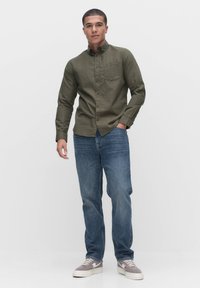 Olijfgroene overhemdd met lange mouwen, borstzak en zichtbare knopen, gecombineerd met blauwe denim jeans en lichtgrijze sneakers.
