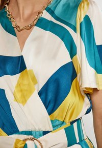 Lyhythihainen kietaisumekko silkkisessä kankaassa, jossa on sinisiä, teal-värisiä ja keltaisia abstrakteja kuvioita. Mekossa on rypytetty vyötärö ja kultaketjuvyö-yksityiskohta.