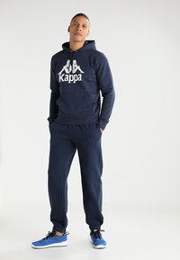 Tumši zilā Kappa treniņtērps ietver kapuci ar lielu baltu logo, saskaņotas bikses un zilus sporta apavus ar melniem akcentiem.
