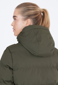 Chaqueta acolchada verde oliva con capucha, que presenta un diseño acolchado y una textura suave. La vista trasera destaca la forma y las costuras de la capucha.