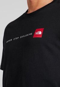 Černé bavlněné tričko s klasickým kulatým výstřihem, s textem "NEVER STOP EXPLORING" a červeným čtvercovým logem značky The North Face na prsou.