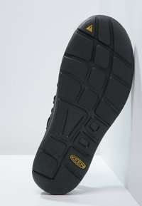 De zwarte rubberen schoenzool heeft diepe profielgroeven, een verhoogde hiel en een geel accent. De textuur is glad met duidelijke groeven voor grip.