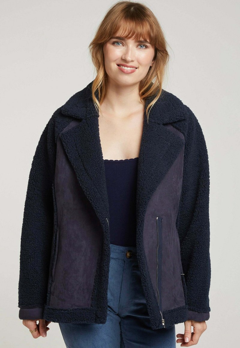 Manteau bleu marine avec un extérieur texturé et duveteux et un tissu intérieur lisse, doté d'un col à revers et de poches avant zippées.