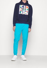 Mörkblå hoodie med färgglad grafik "Polo Ralph Lauren", kombinerad med ljusblåa sweatpants. Vita sneakers fullbordar outfiten.