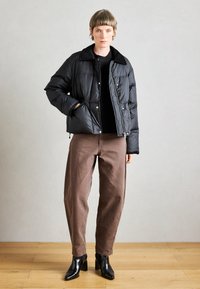 Črna napihnjena jakna s ovratnikom in zapenjanjem na gumbe, v kombinaciji s rjavimi hlačami in črnimi škornji s peto, na nevtralni podlagi.