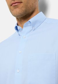 Camicia azzurro chiaro con colletto, dotata di taschino applicato e bottoni bianchi. La texture sembra liscia e tessuta.